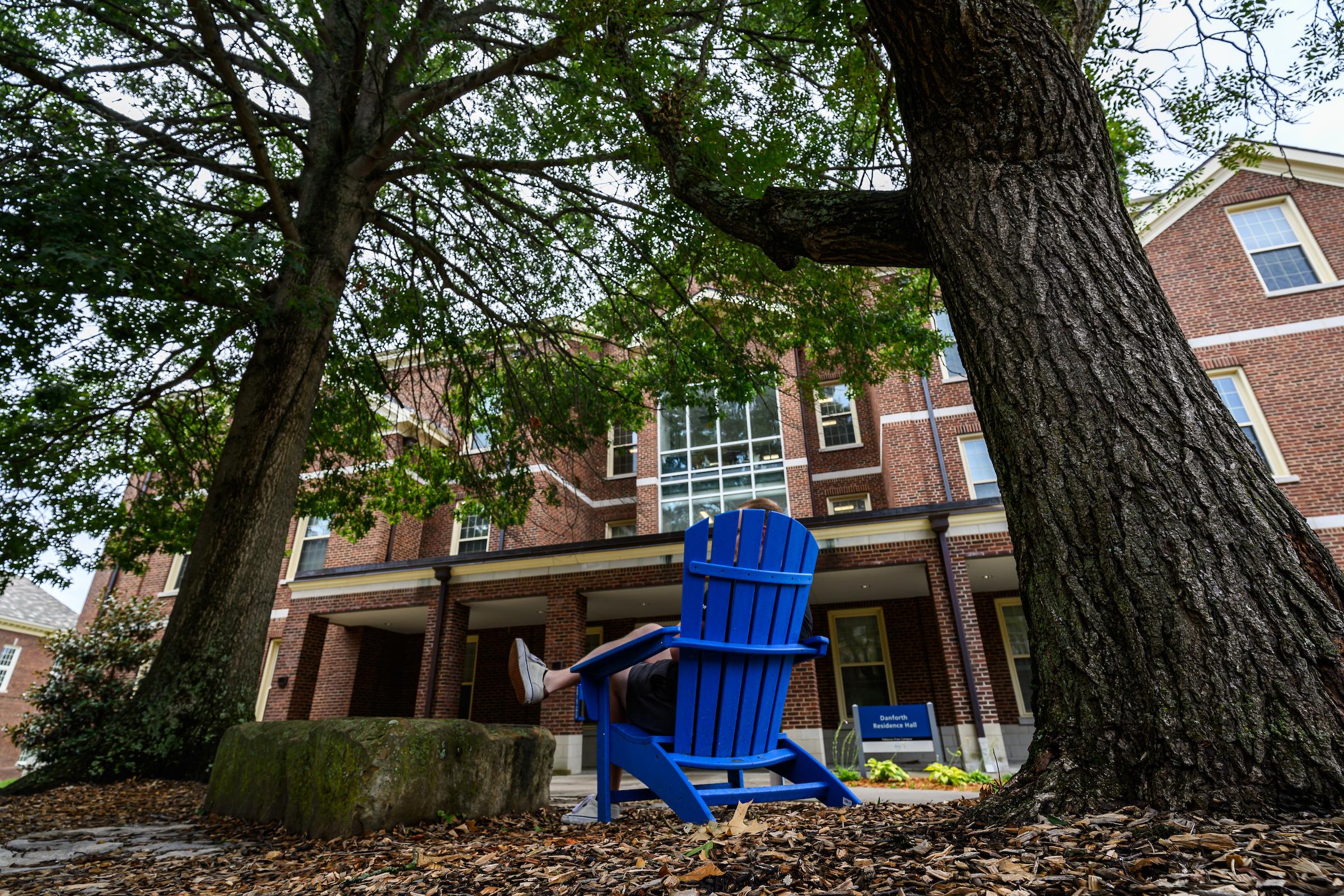 The back of blue chair in front of one of the student dorms. A person's leg can be seen, crossed.
